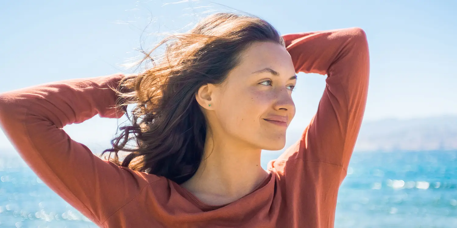  Junge Frau mit langen braunen Haaren blickt lächelnd zur Seite am Meer