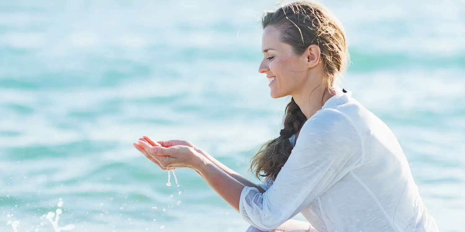  Frau mit Zopf schöpft lächelnd Wasser am Meer mit beiden Händen