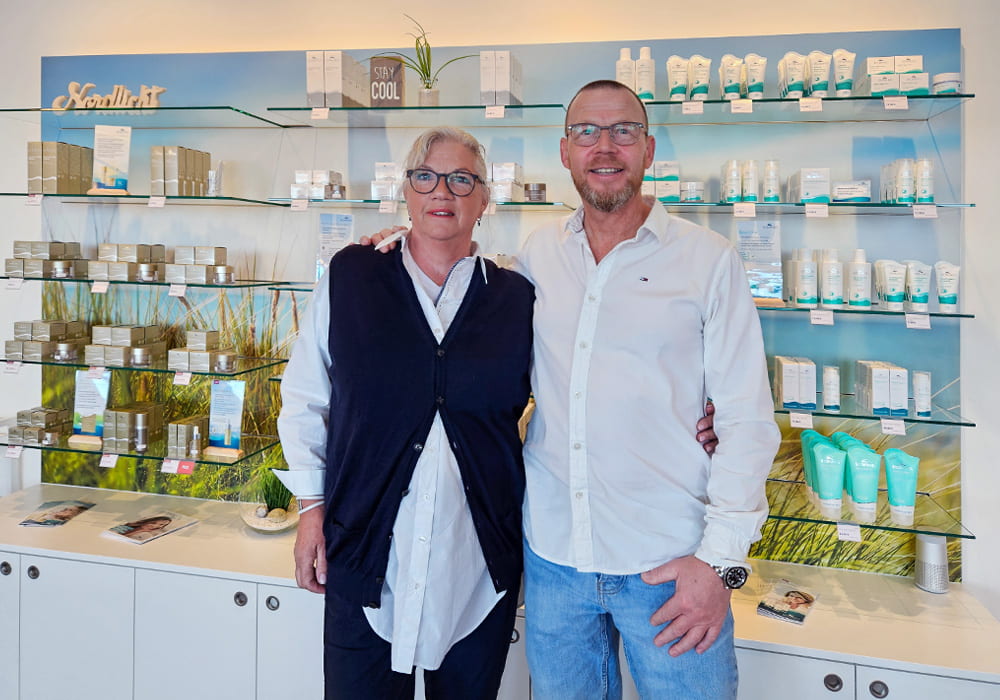 Ein Shop-Betreiber und eine Mitarbeiterin stehen  lächelnd vor einem Verkaufsregal mit Biomaris-Produkten im modern gestalteten Laden.