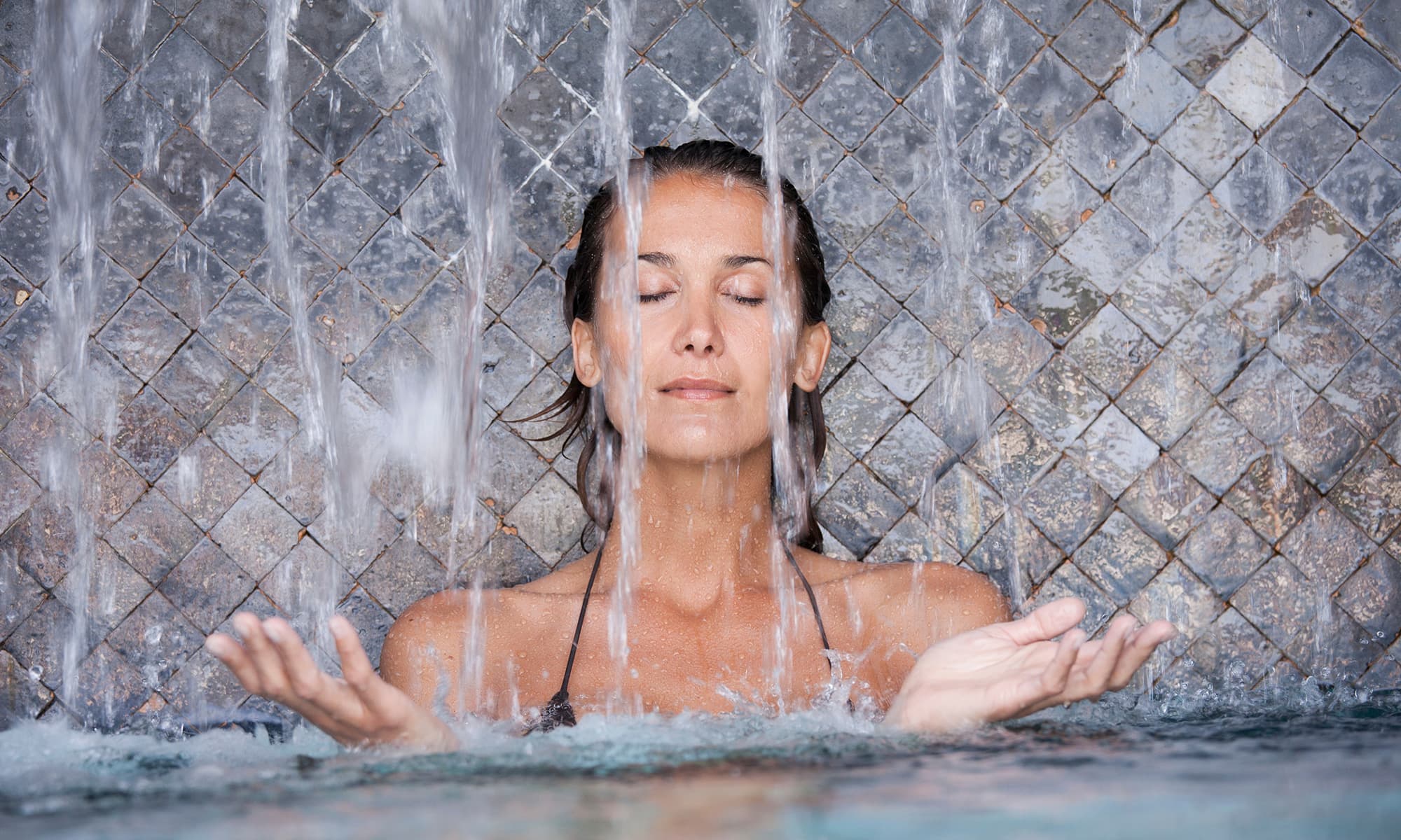  Frau mit geschlossenen Augen genießt einen Wasserfall in einem Spa-Bereich vor gefliestem Hintergrund.
