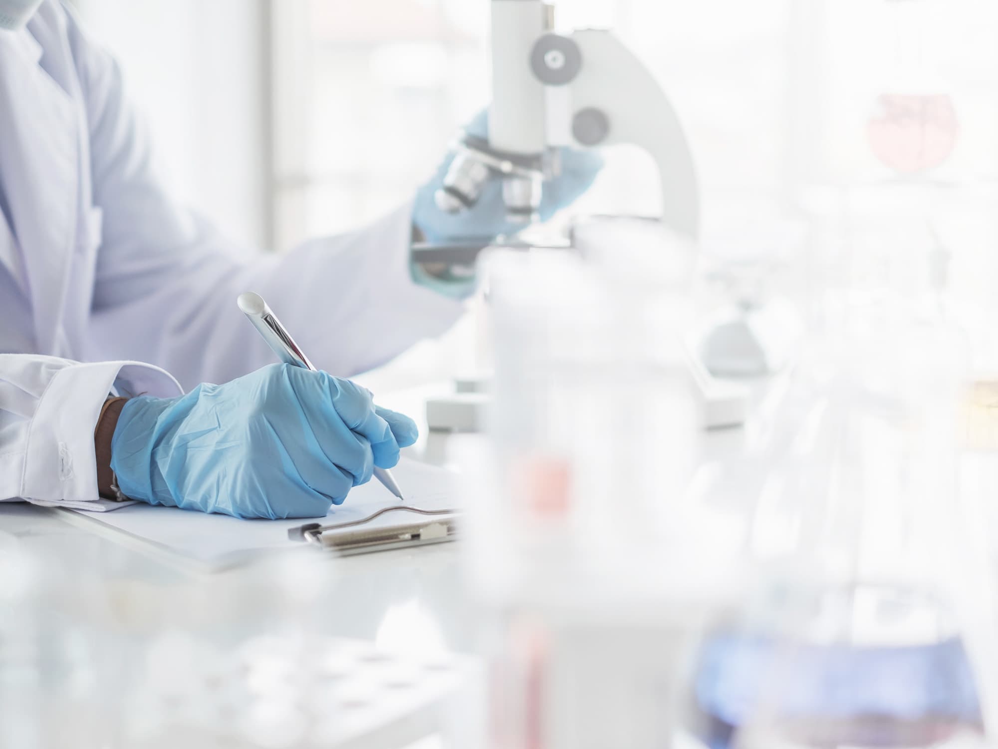  	Wissenschaftler mit blauen Handschuhen hält einen Stift und schreibt auf einem Klemmbrett, während er ein Mikroskop betrachtet.